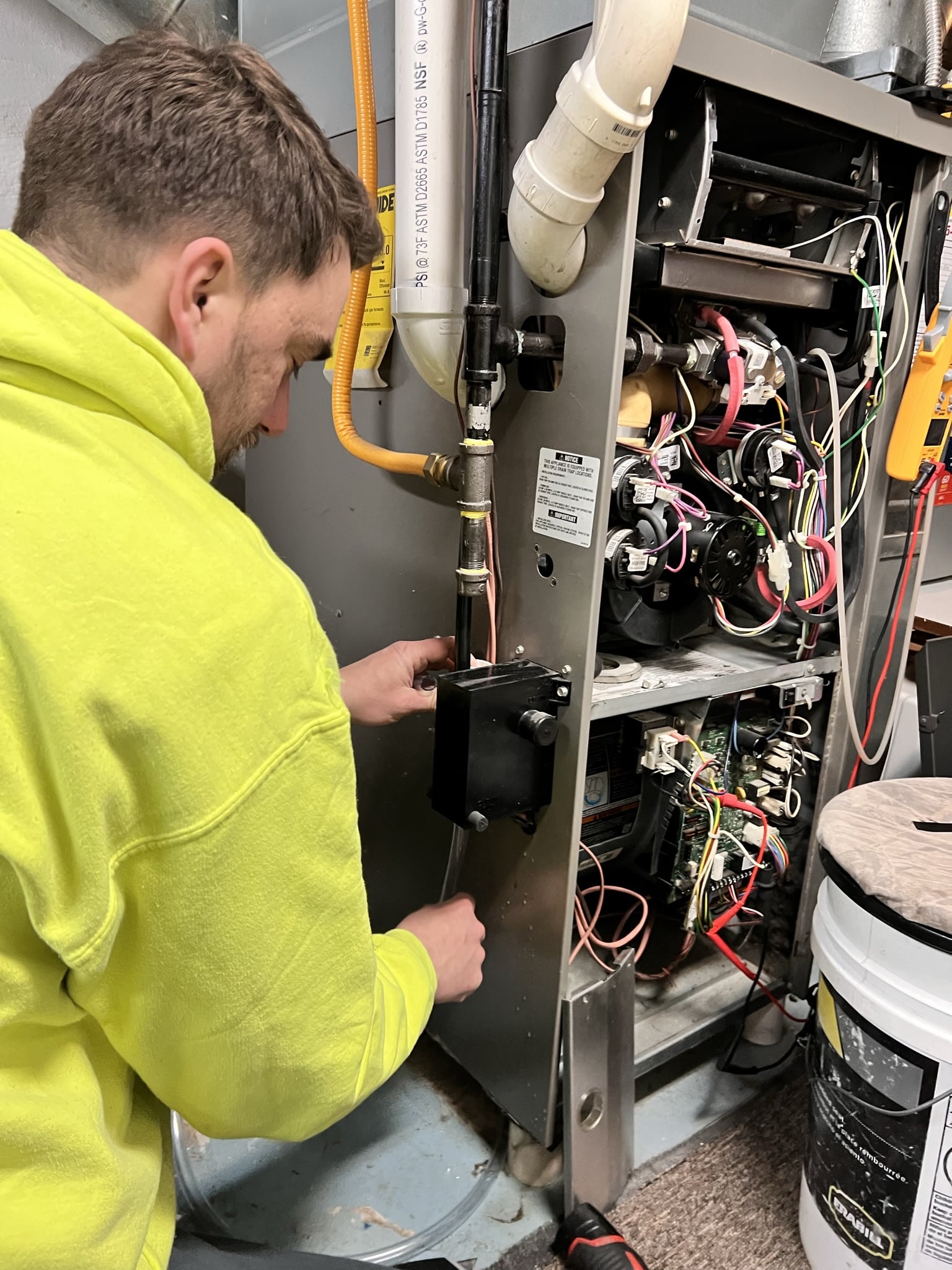 A person in a yellow jacket repairs a complex metal furnace unit with colorful wires, pipes, and valves, concentrating on the task.
