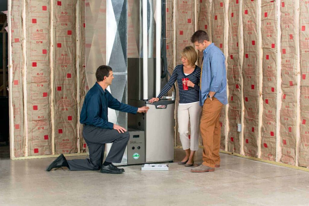 Three people inspect a furnace in an unfinished room with exposed insulation. One person kneels and gestures toward the appliance controls.