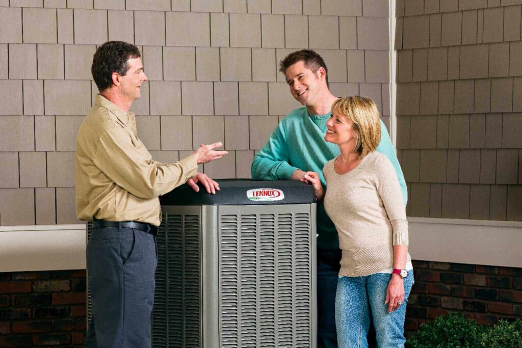 Three people stand around a Lennox air conditioning unit, engaging in conversation, outside a house with brown siding and brick accents.