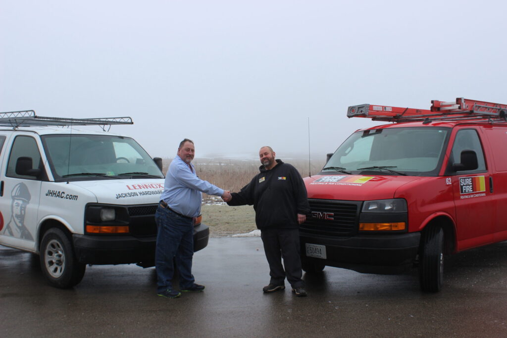 Two persons shake hands between parked service vans labeled "Jackson Hardware" and "Sure Fire," on a misty day. No landmarks visible.