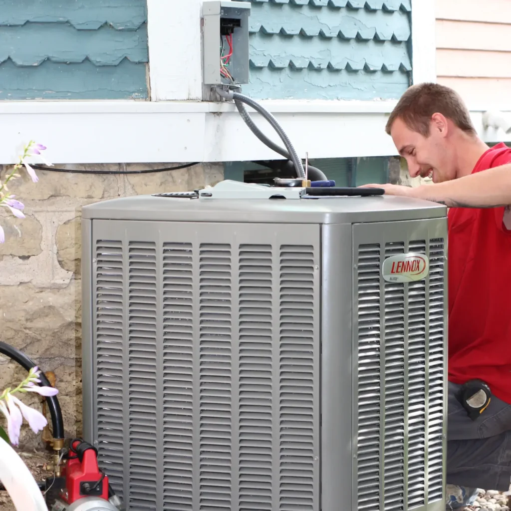 A person in a red shirt services a Lennox air conditioning unit outside a house with blue and white siding.