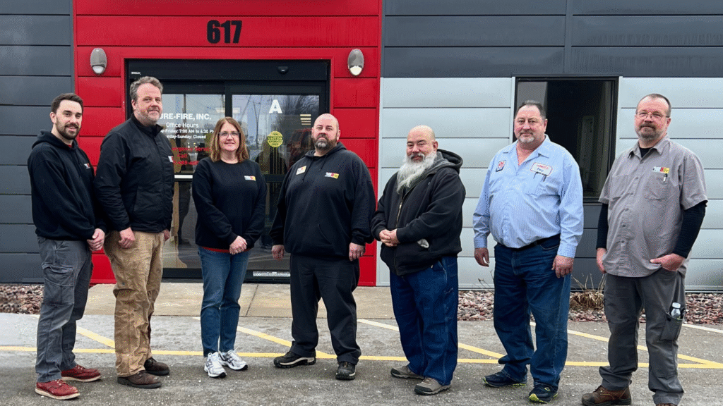 Seven people stand outside a building with a red and gray facade, labeled 617. They are casually dressed and smiling.