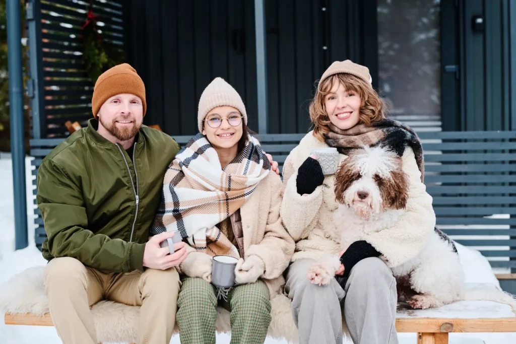 Three people seated outside in winter attire, holding mugs, with a fluffy dog beside them. Snow is visible. Cozy and cheerful atmosphere.