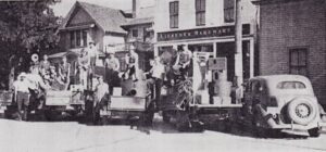 A group of people on trucks in front of Lifesner Hardware store, with a vintage car parked nearby on a sunny day.