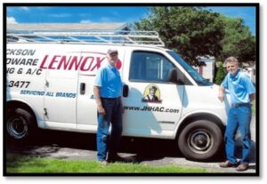 Two people stand next to a van labeled "Lennox," parked on a driveway. Both wear blue shirts, with trees in the background.